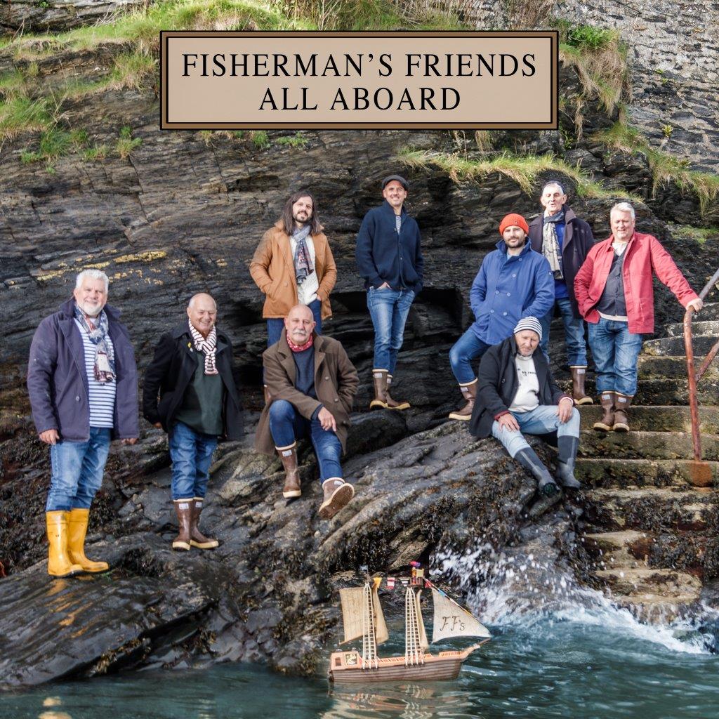 Fisherman’s Friends band members pose with a model sailboat by rocky coastline.