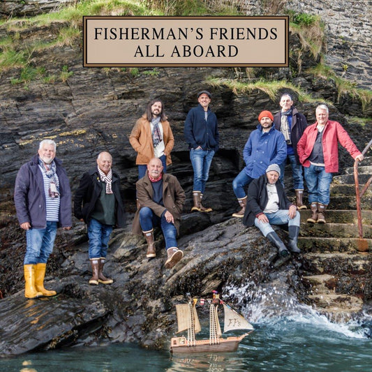 Fisherman’s Friends band members pose with a model sailboat by rocky coastline.