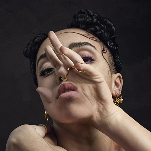 Fka Twigs portrait with gold jewelry, hand partially covering face.