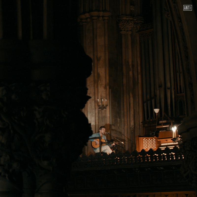 A man plays acoustic guitar in a dimly lit, vaulted stone church interior.