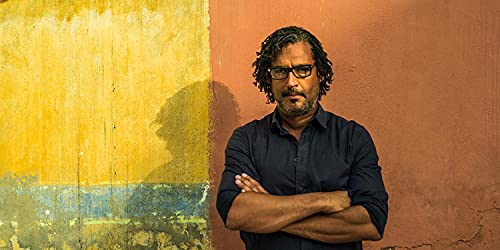 A man with glasses and dreadlocks stands with arms crossed against a colorful wall.