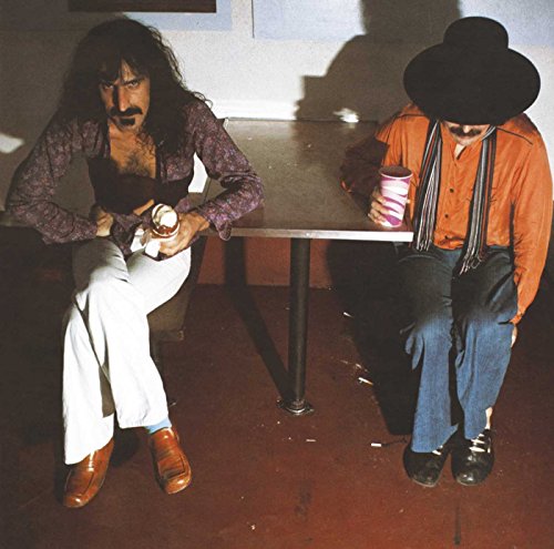 Frank Zappa and Captain Beefheart sit at a table in a dimly lit room.