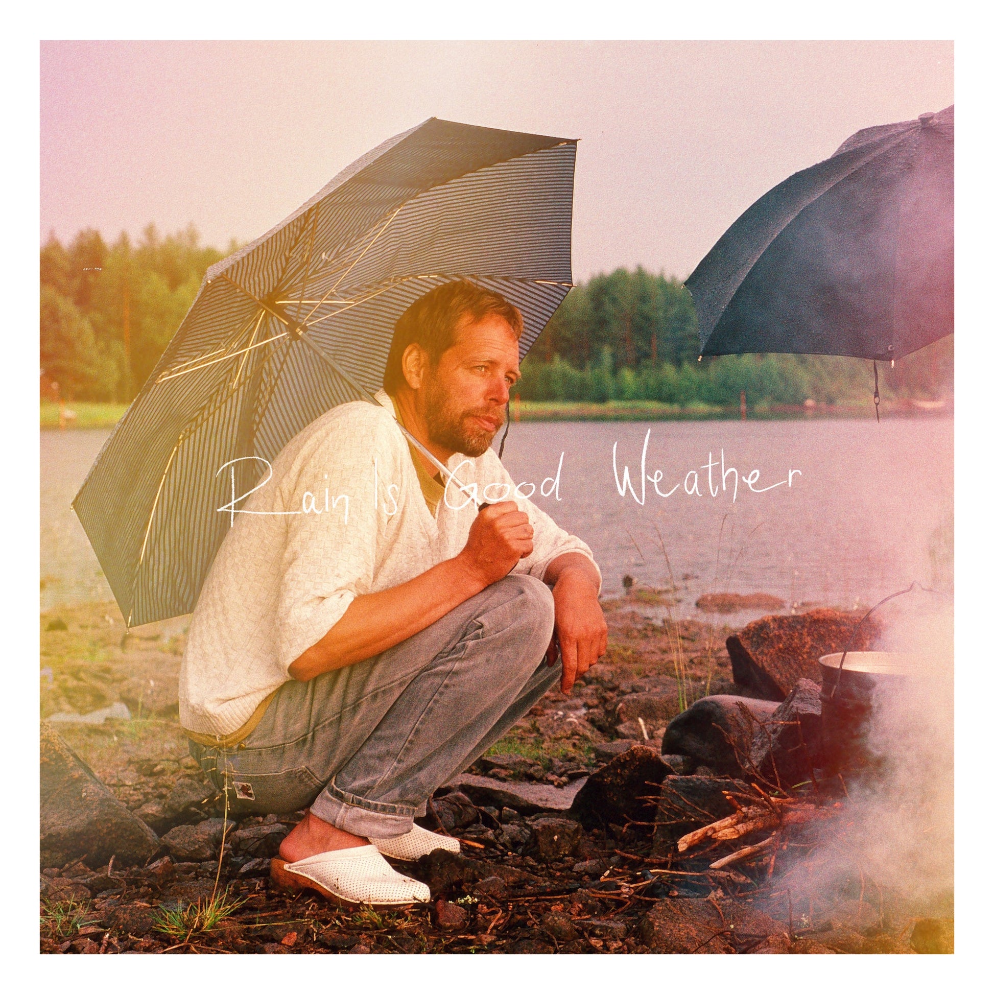 A man crouches under an umbrella by a lake near a smoking campfire.