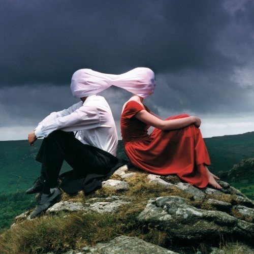 A man and woman sit on a rock, heads covered by a single white cloth.