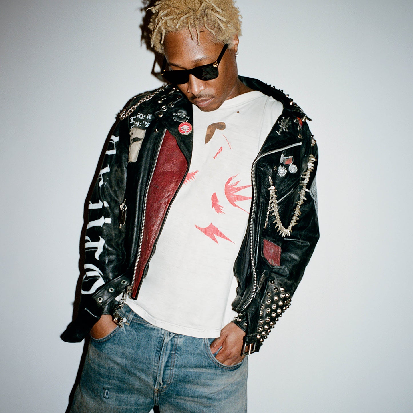 Man with blonde curly hair, sunglasses, and a studded leather jacket against white background.