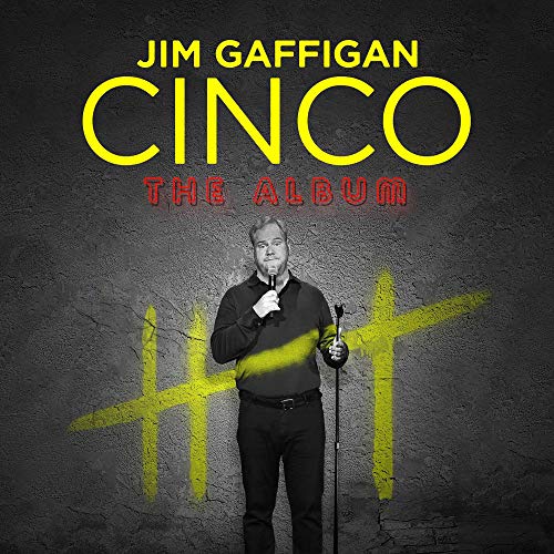 Jim Gaffigan stands holding a microphone in front of a wall with yellow tally marks.