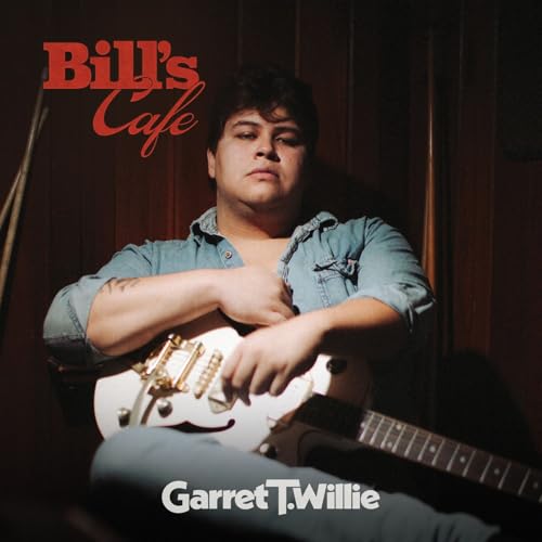 Garret T. Willie sits in front of a wood wall holding an electric guitar.