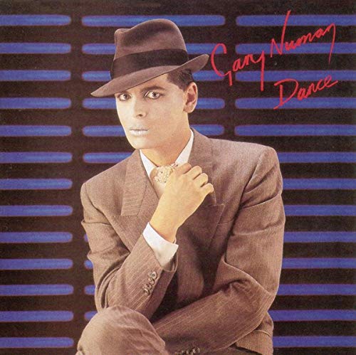 Gary Numan wearing a fedora and brown suit against a blue striped background.