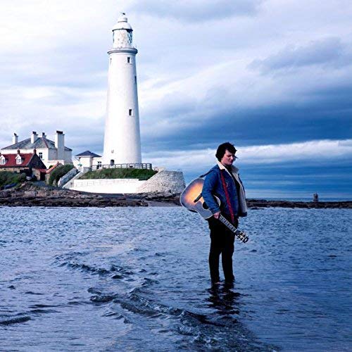 Man with guitar stands in water before a lighthouse and buildings.