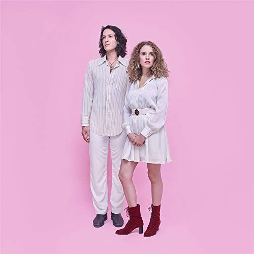 "Man and woman in white clothing standing against a pink background."