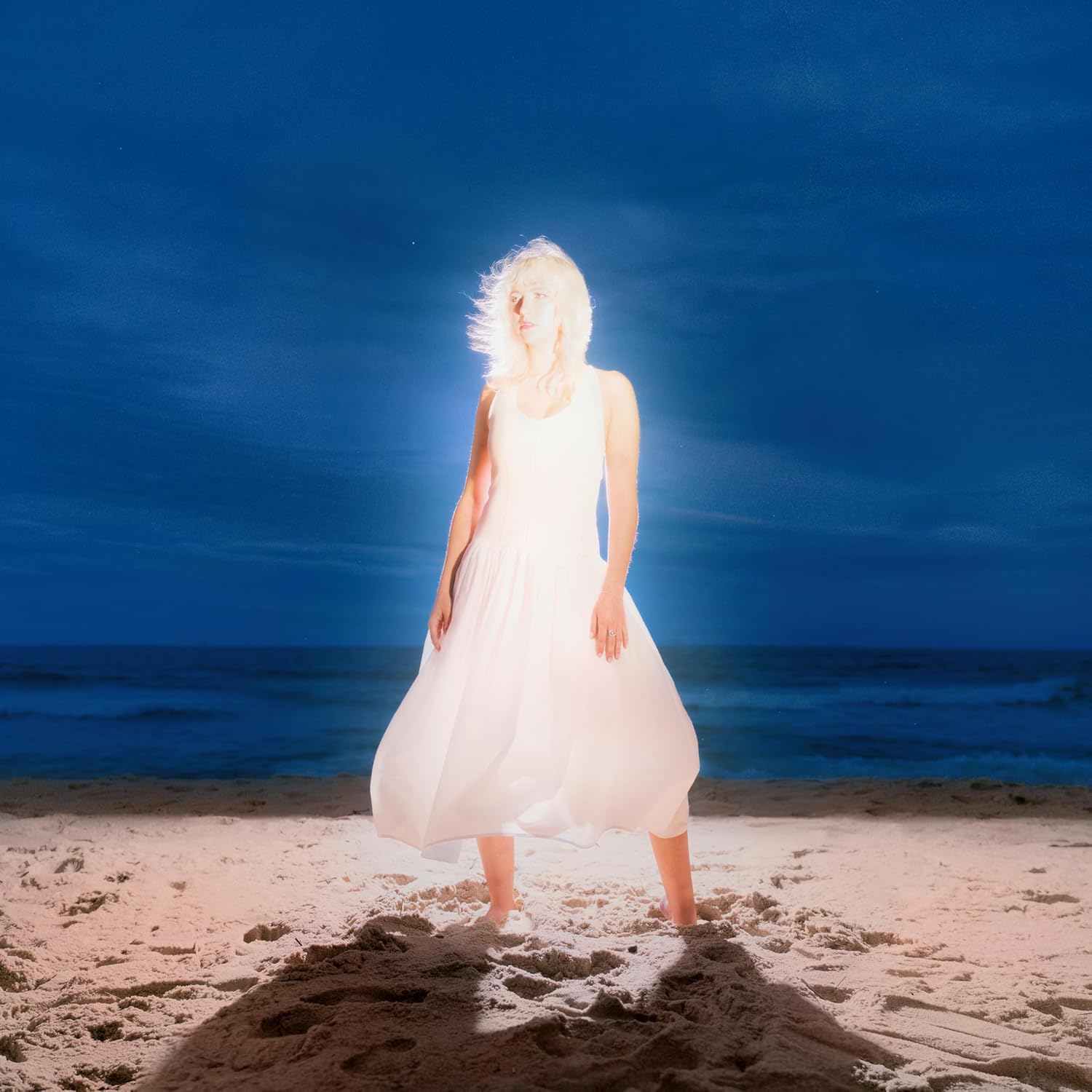 Woman in white dress stands on a beach, backlit by bright light.