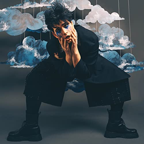 Man with painted tears poses dramatically amidst hanging blue clouds.