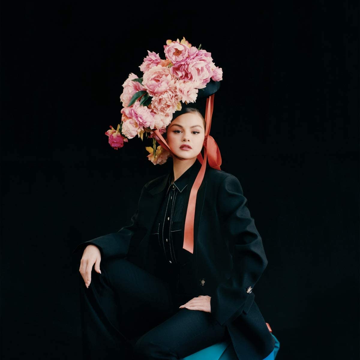 Woman in black outfit, wearing a large floral headpiece, against a dark background.