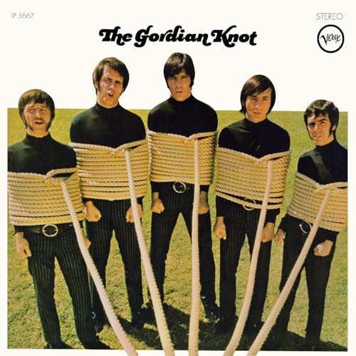 Four men in black turtlenecks tied with rope stand in a grassy field.