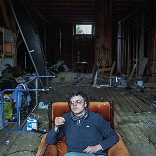 Man with glasses sits in an orange armchair in a dilapidated room.