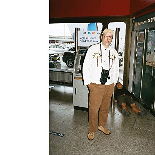 A man stands in a bank, wearing a hat, glasses, and a camera around his neck.