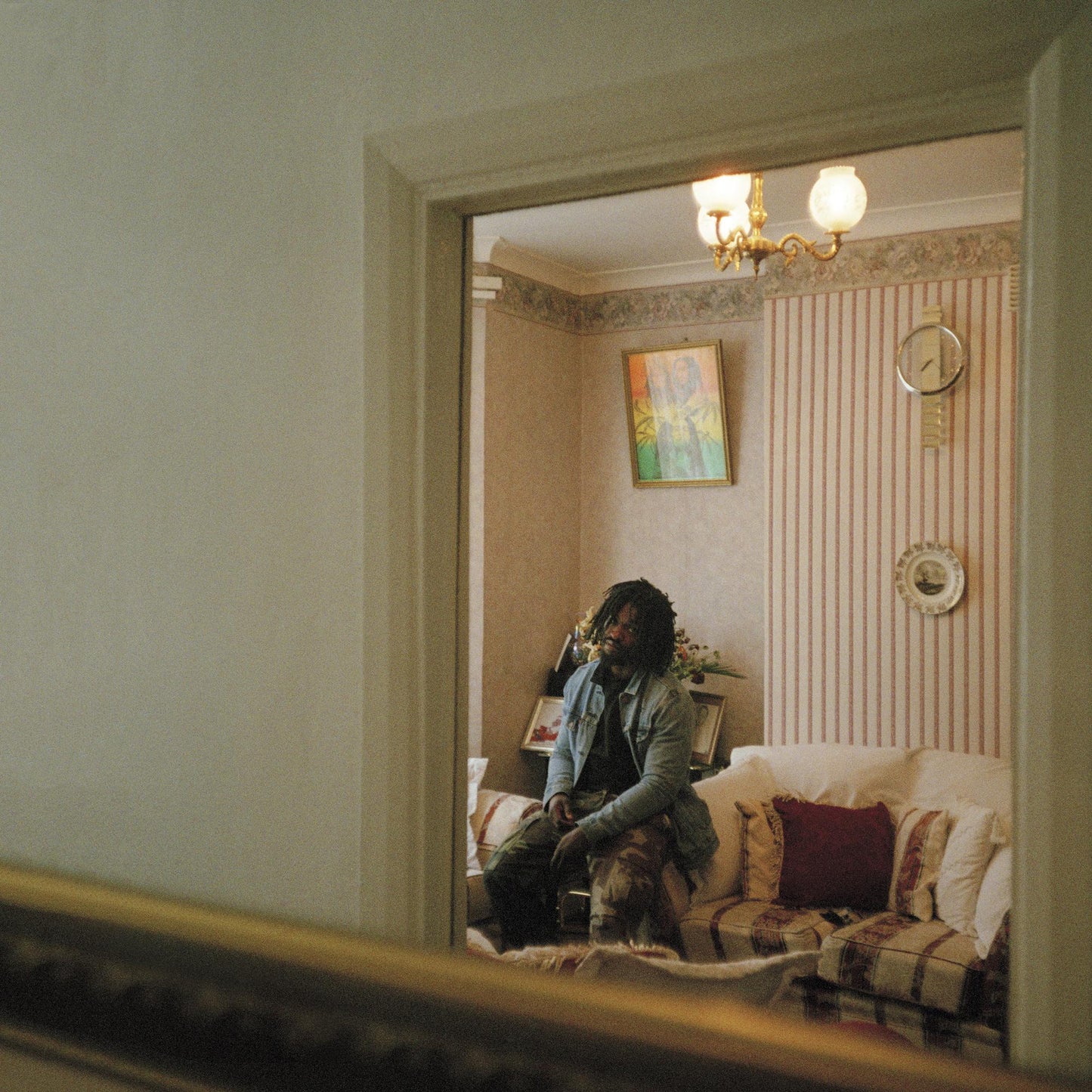Man with dreadlocks sits on a sofa in a patterned room.