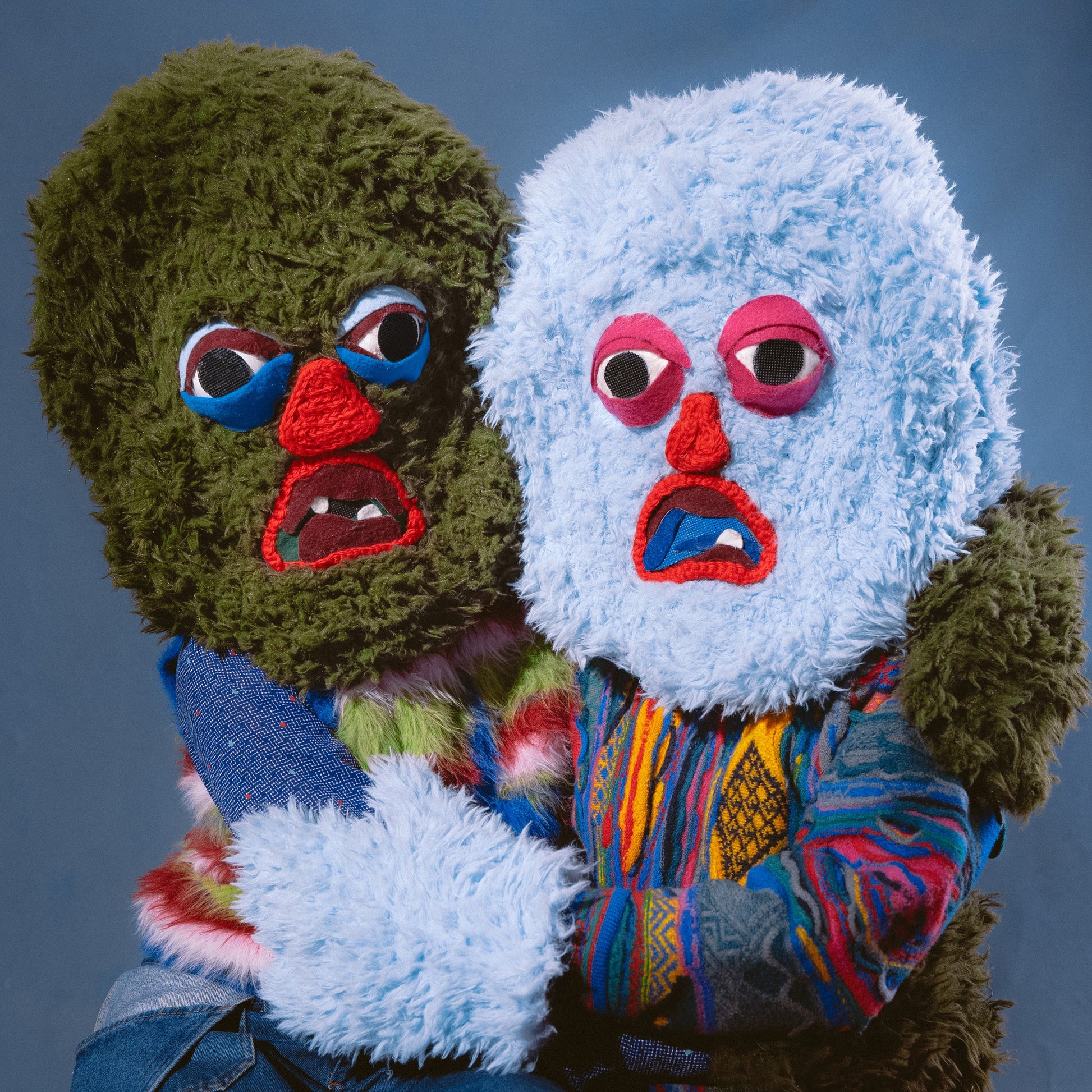 Two people wearing fuzzy green and blue monster masks and colorful patterned sweaters.