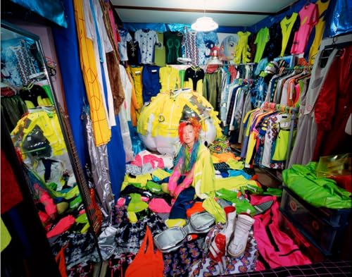 A person with bright pink hair sits surrounded by colorful clothes in a cluttered room.