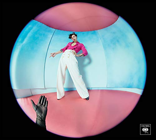 Harry Styles poses in a pink and blue circular room, hand reaching up.
