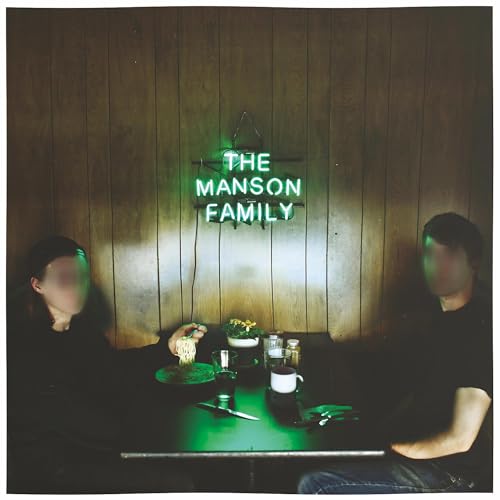 Two people with blurred faces sit at a table beneath a green neon sign.