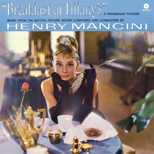 Audrey Hepburn sits at a table in a black dress, pearls, and long gloves.