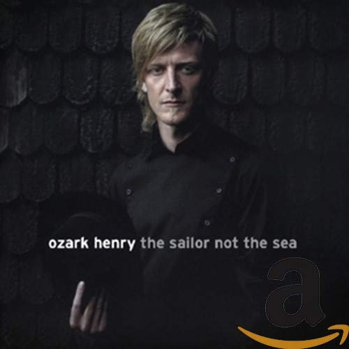 A man with blonde hair in a black shirt holding a hat against dark shingles.