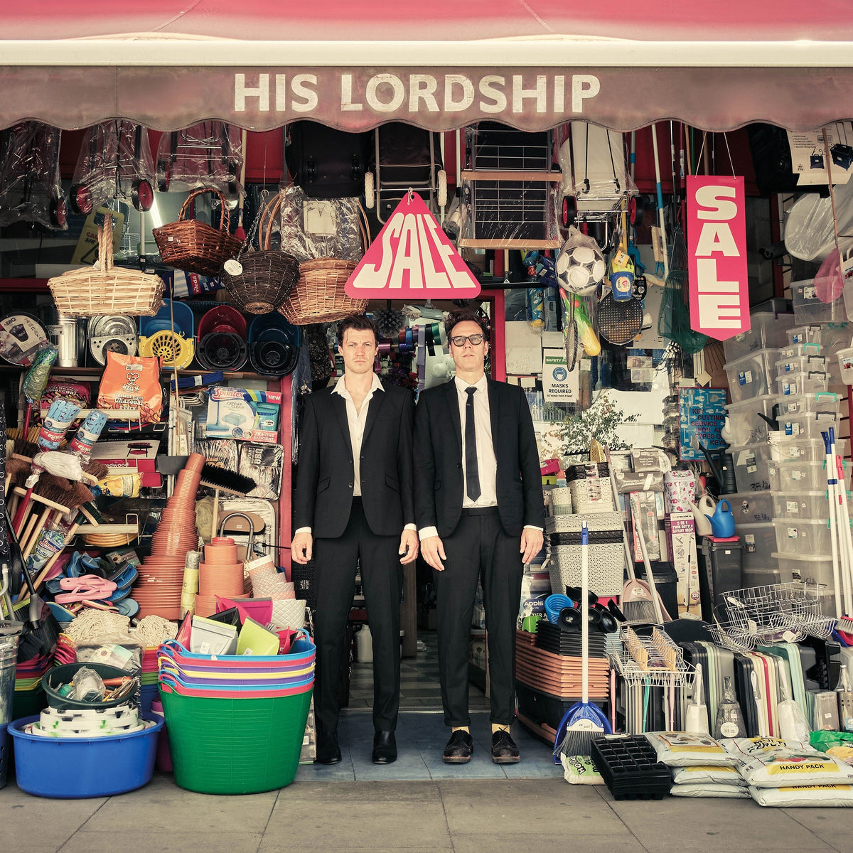 Two men in suits stand before a cluttered market stall display.