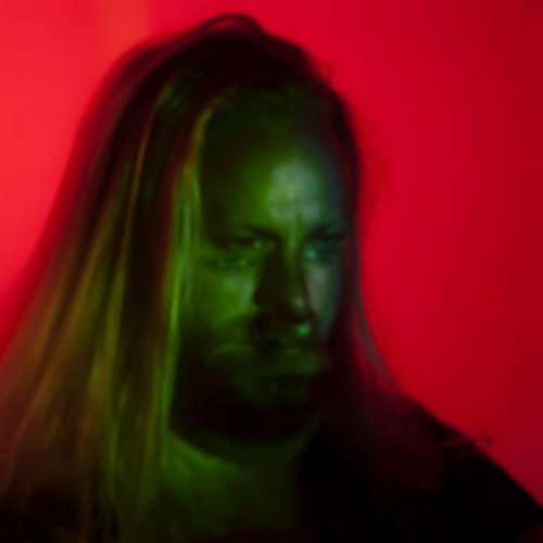 Close-up portrait of a man with long hair, lit with red and green tones.