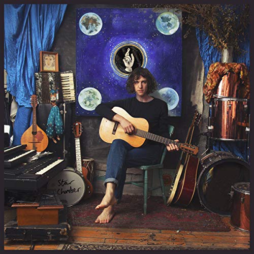 Man sits on stool, holding guitar, surrounded by musical instruments and celestial-themed tapestry.