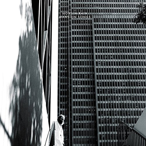Black and white photo of man standing beside a building with skyscraper behind.