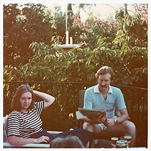 A man and woman sit outdoors on a patio with foliage in the background.