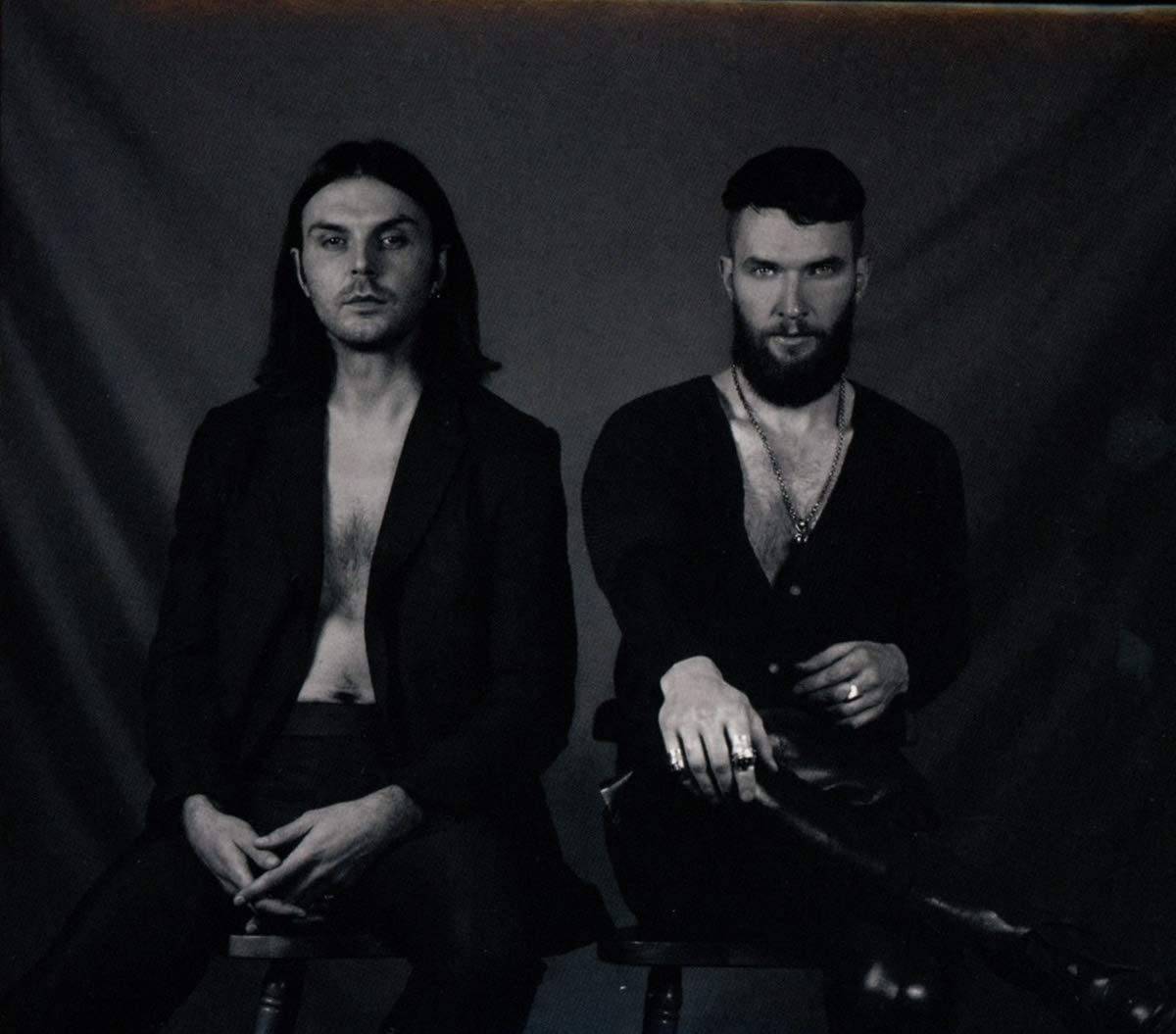 Black and white image of two men sitting on stools, dressed in dark clothing.