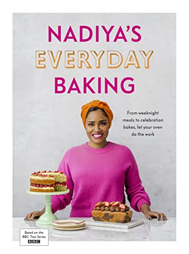 Nadiya Hussain wearing an orange headwrap and pink sweater behind two baked cakes.