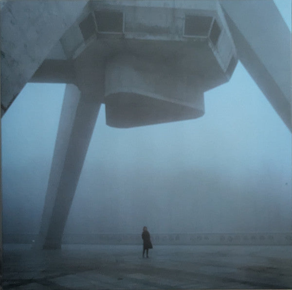 A lone figure stands under a large, concrete structure in dense fog.