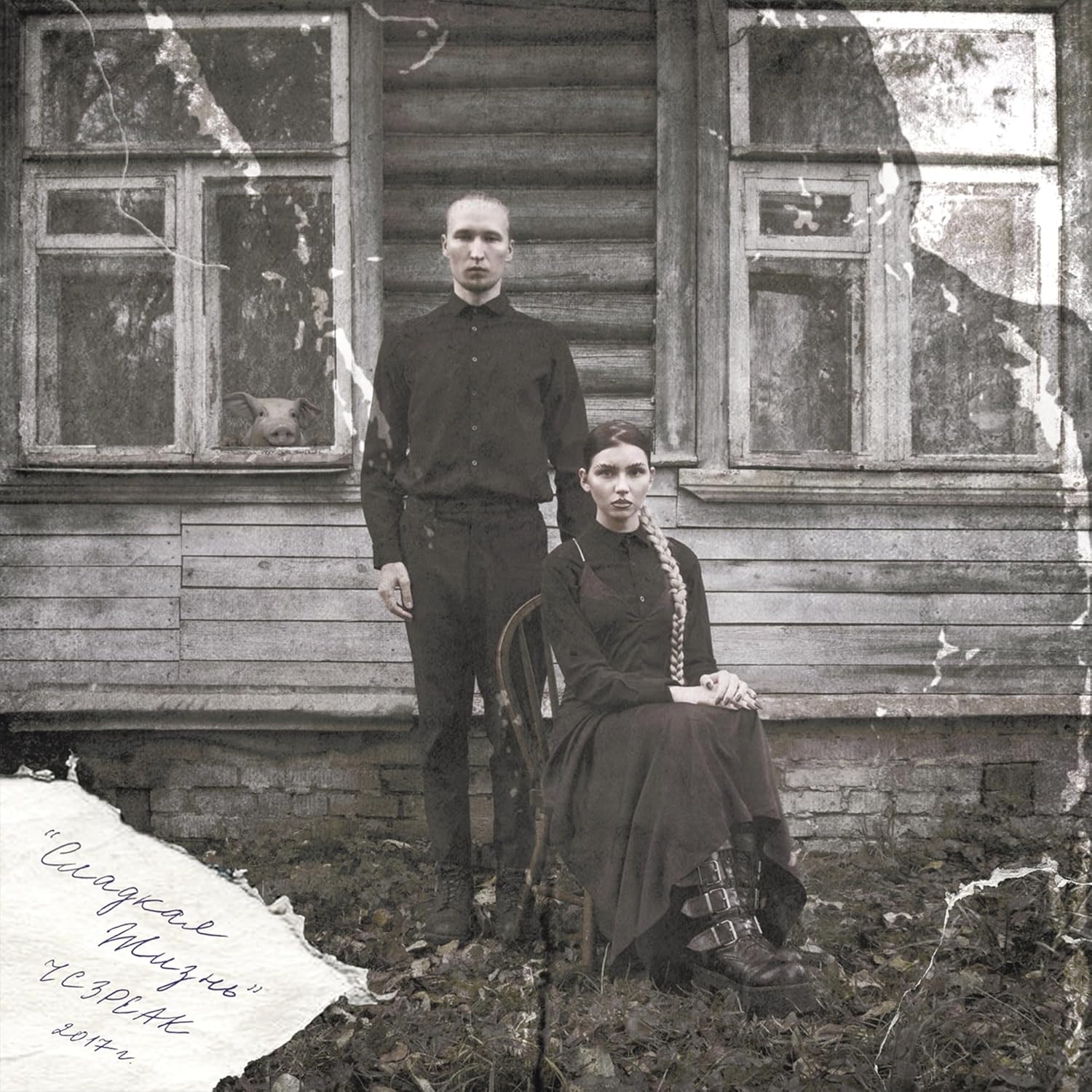 Man and woman in black clothing posing before a weathered wooden house.