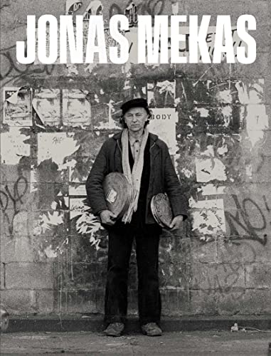 Black and white portrait of Jonas Mekas standing before a graffitied wall.