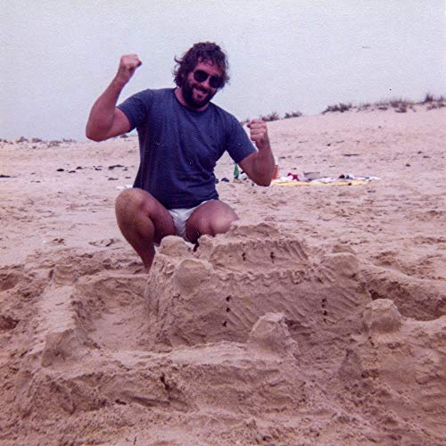 Man crouching by a sandcastle on a beach, raising fists.