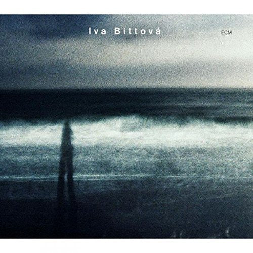 "Album cover featuring a person standing on a beach with a dark, cloudy sky."