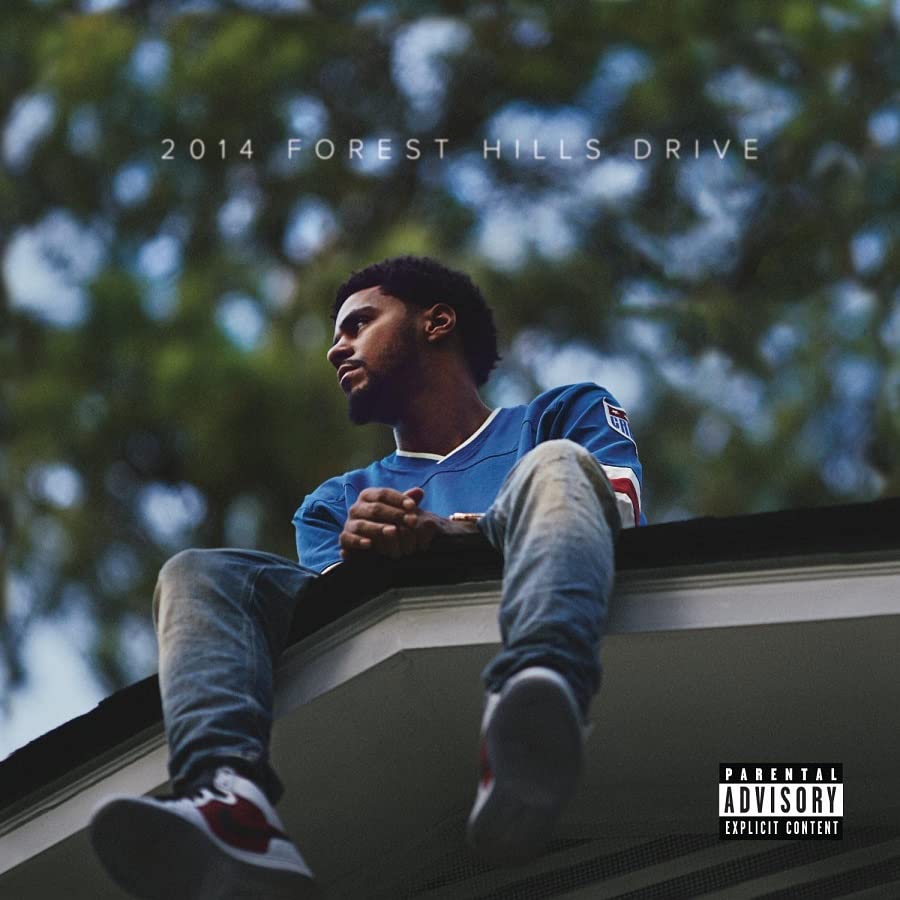 J. Cole sitting on a roof against a blurred background of trees.