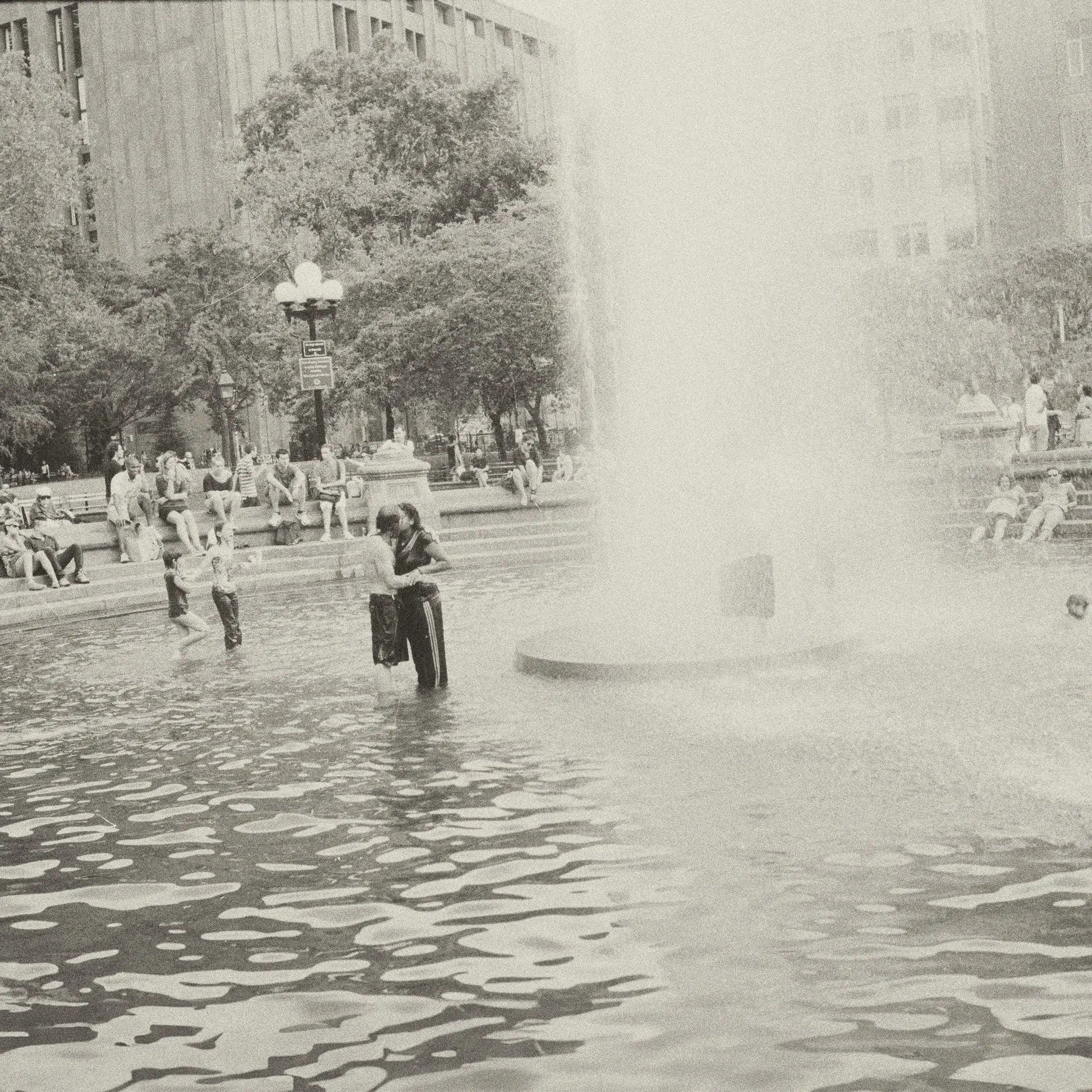 Black and white photo of people in a city park fountain with a large spray.