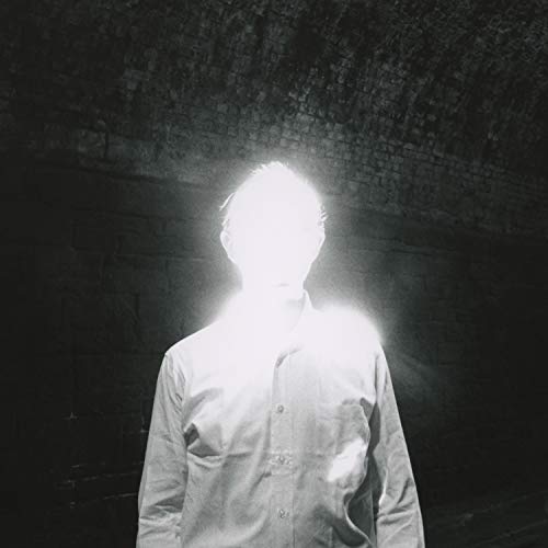 Man in a white shirt, overexposed, standing in a dark brick-walled room.