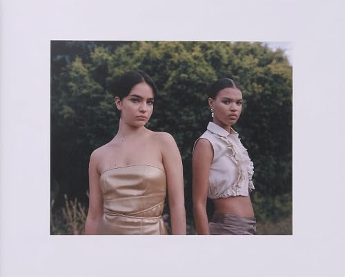 Two young women with serious expressions, partially exposed shoulders, and greenery backdrop.