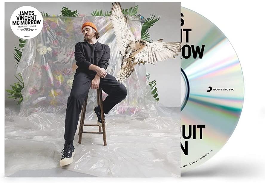 Man in black outfit sits on stool with owl, surrounded by flowers and plastic sheeting.