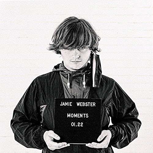Black and white portrait of Jamie Webster holding a sign reading "Moments.