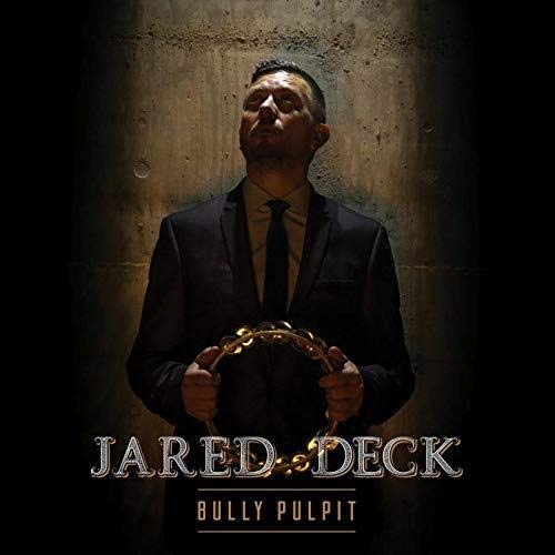 Jared Deck portrait, holding a tambourine, against a dark concrete wall.