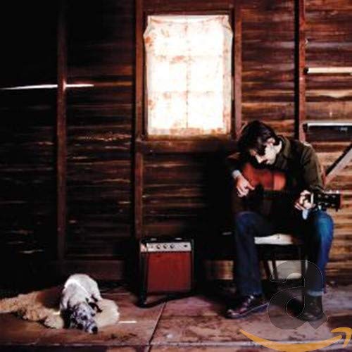 Man with guitar seated in front of a rustic wooden building.