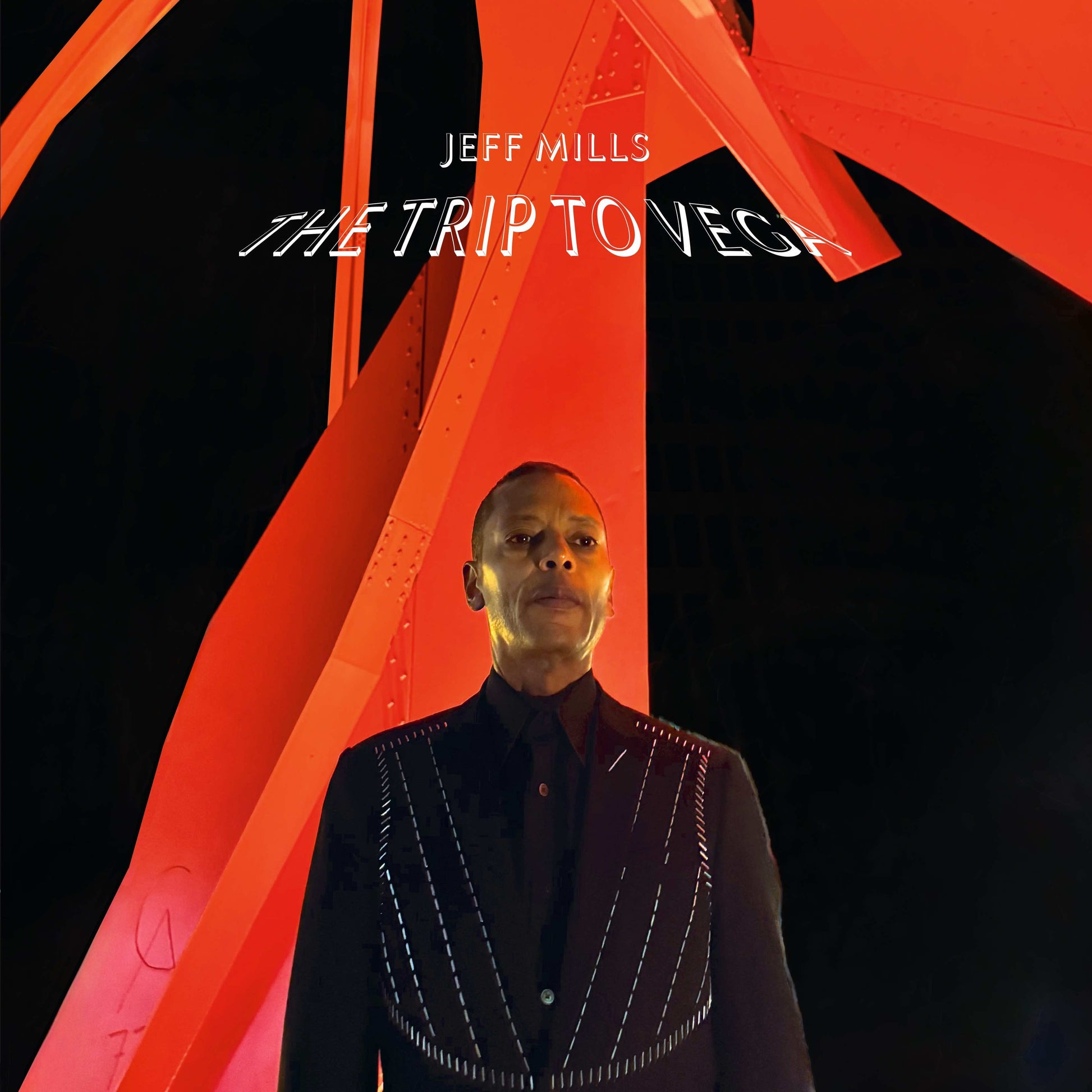 Jeff Mills in a black suit standing before a large, bright red abstract structure.