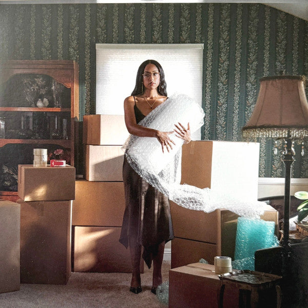 Woman in slip dress holding bubble wrap, surrounded by cardboard boxes.