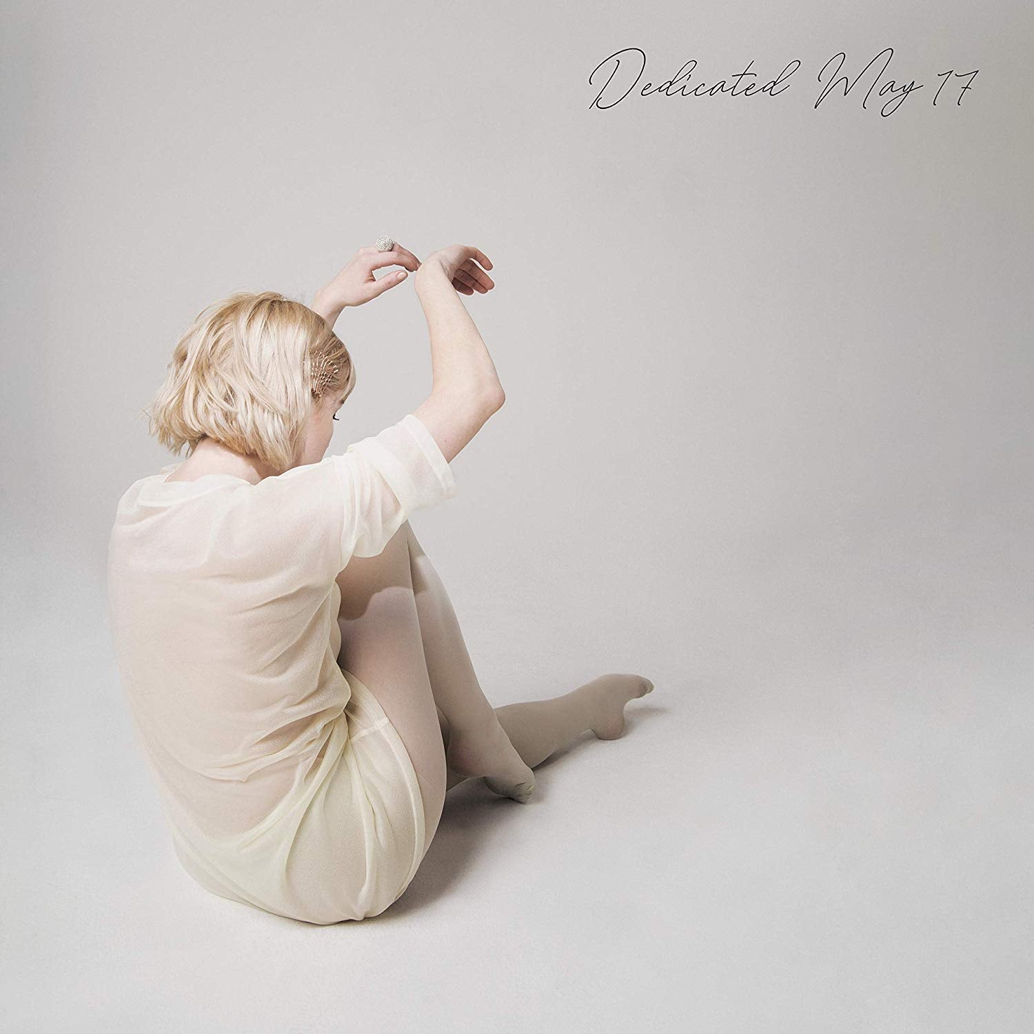 Woman in white fringe pants, bare back, and short hair, standing against a beige background.